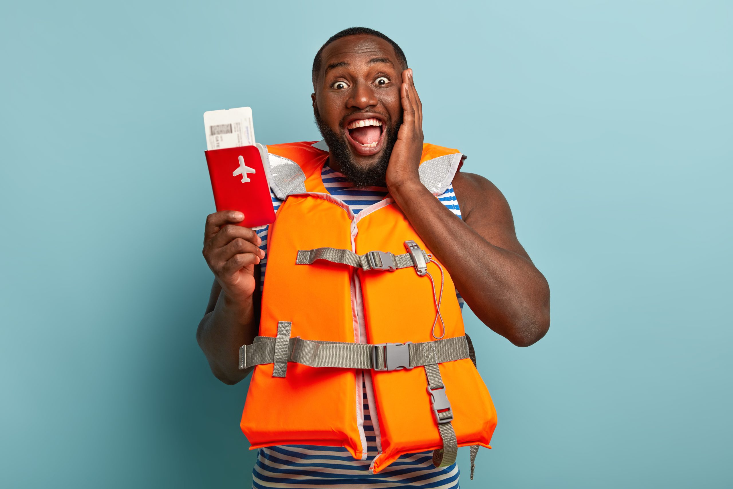 Half length shot of happy black man enjoys coming travel, holds red passport with tickets, shares impressions, wears lifejacket, touches cheek, isolated on blue studio wall. Summer time and tourism