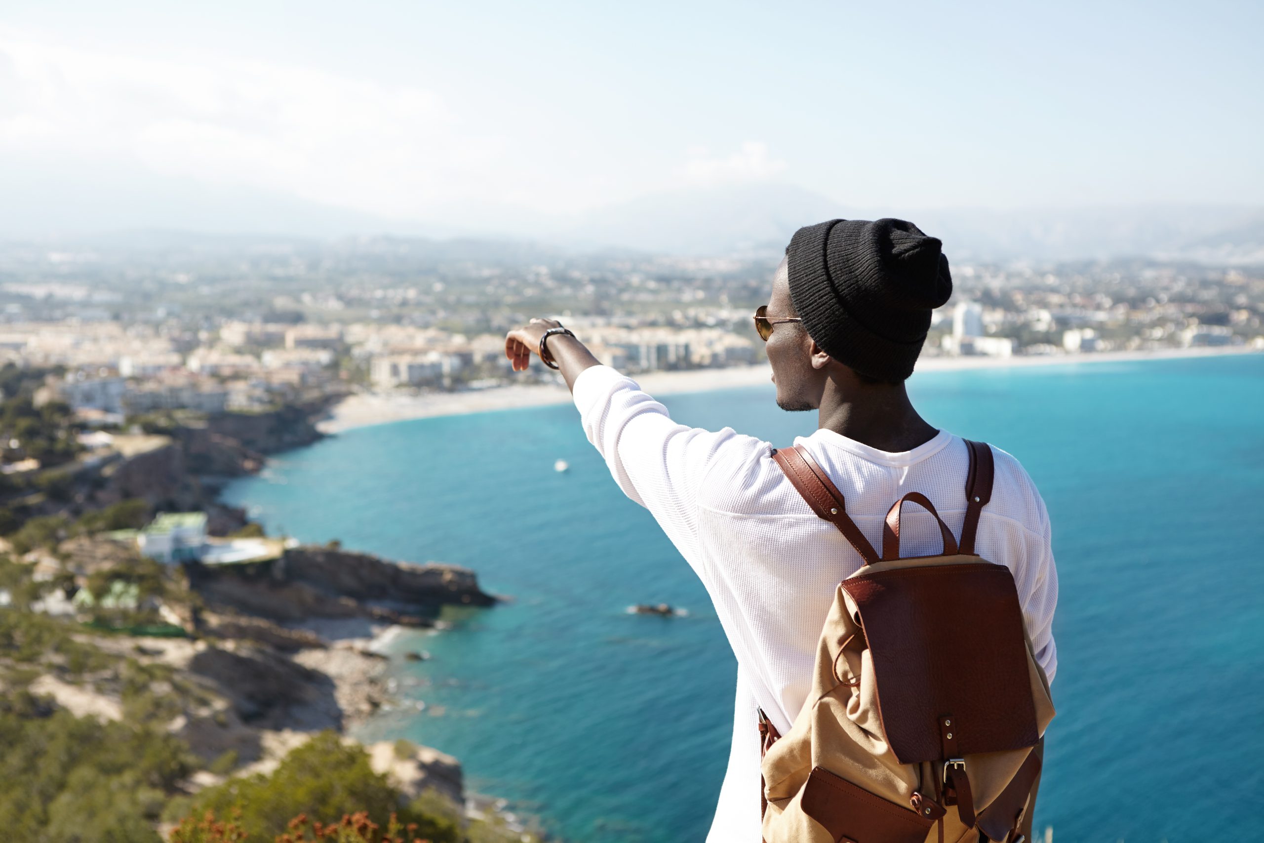 Portrait of unrecognizable young African man in black hipster hat standing on sightseeing platform admiring sea and beautiful resort town pointing his finger at faraway places he's going to visit