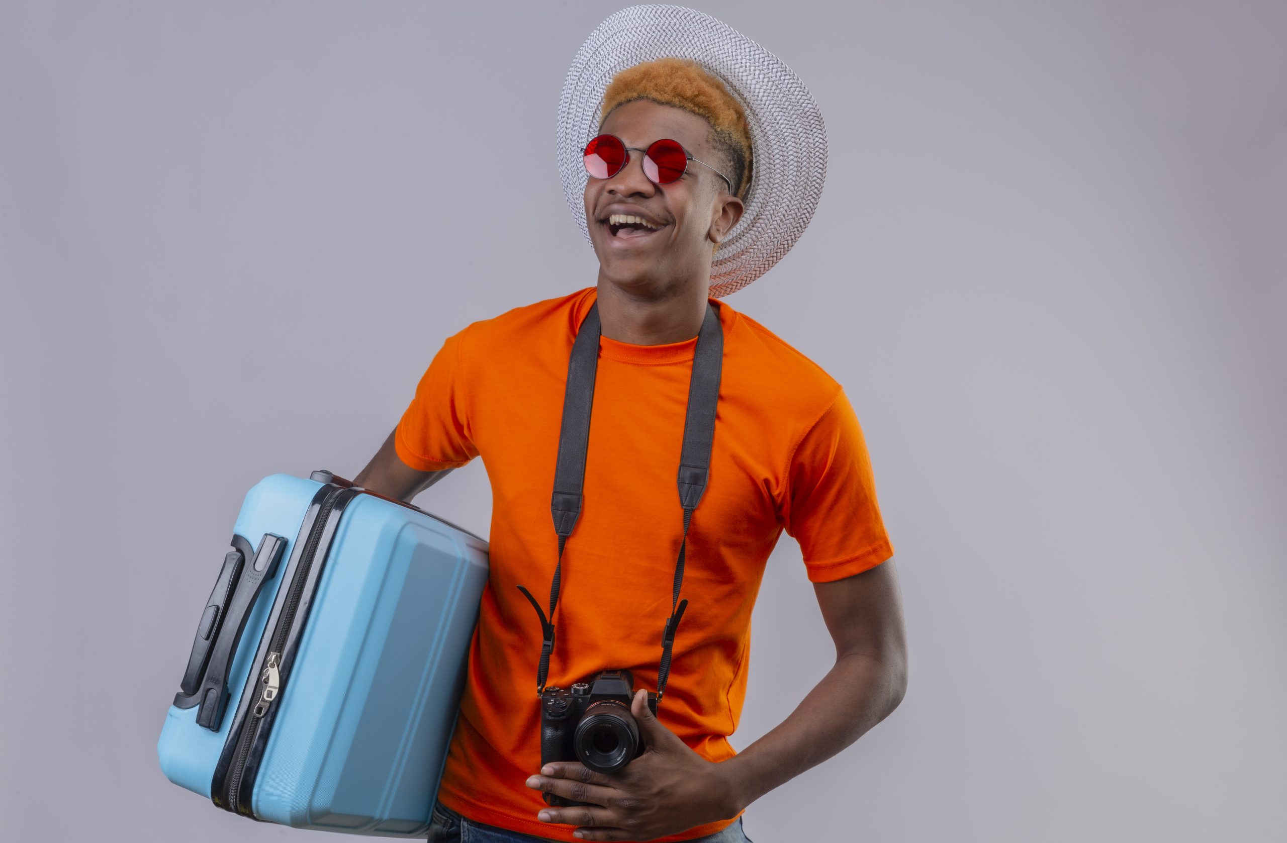 young african american handsome traveler boy in summer hat wearing orange t-shirt holding travel suitcase laughing happy and positive standing over white background